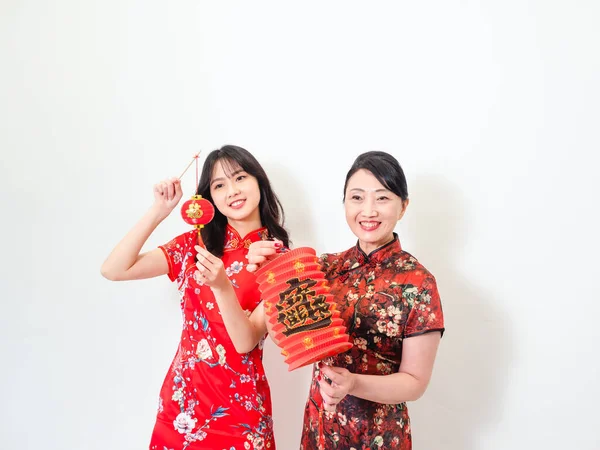 Portrait of young asian woman and elder woman both wearing traditional cheongsam qipao dress hold red paper lantern with word meaning good fortune and treasure on white background.