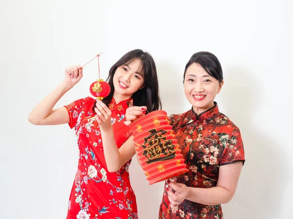 Portrait of young asian woman and elder woman both wearing traditional cheongsam qipao dress hold red paper lantern with word meaning good fortune and treasure on white background.