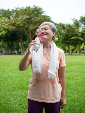 60 'ların yaşlı, yaşlı Asyalı kadının portresi ayakta gülümseyen, elinde su şişesi tutan ve parkta temiz havanın tadını çıkaran bir kadın..
