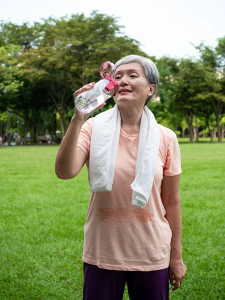 Portrait of 60s senior adult elderly asia woman smiling standing and holding bottled water after sport and enjoy with nature fresh air in the park.