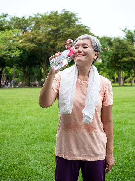 Portrait of 60s senior adult elderly asia woman smiling standing and holding bottled water after sport and enjoy with nature fresh air in the park.