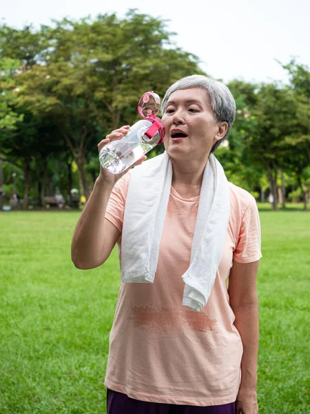 Portrait of 60s senior adult elderly asia woman smiling standing and holding bottled water after sport and enjoy with nature fresh air in the park.