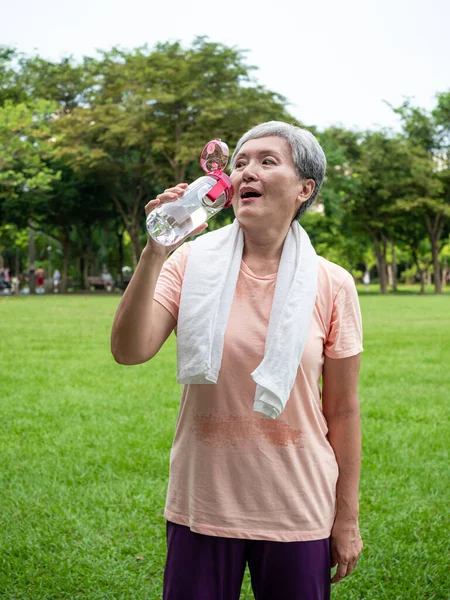 Portrait of 60s senior adult elderly asia woman smiling standing and holding bottled water after sport and enjoy with nature fresh air in the park.