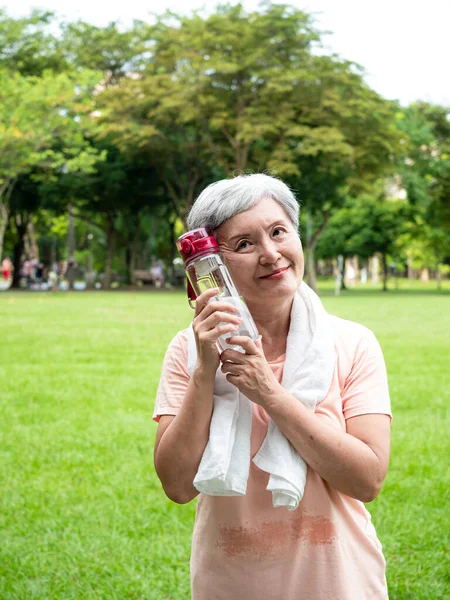 Portrait of 60s senior adult elderly asia woman smiling standing and holding bottled water after sport and enjoy with nature fresh air in the park.