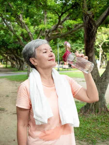 Portrait of 60s senior adult elderly asia woman smiling standing and holding bottled water after sport and enjoy with nature fresh air in the park.