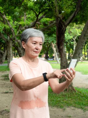 Yaşlı, yaşlı Asyalı bir kadının portresi. 60 'lı yaşlarda, gülümsüyor ve parkta akıllı telefon tutuyor..