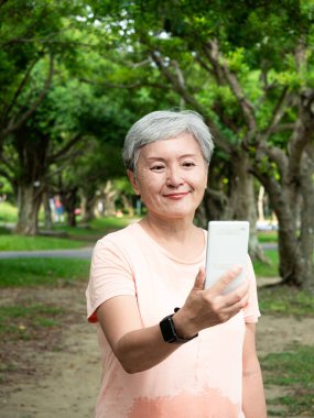 Yaşlı, yaşlı Asyalı bir kadının portresi. 60 'lı yaşlarda, gülümsüyor ve parkta akıllı telefon tutuyor..