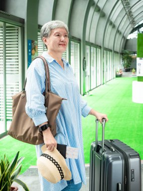 Yaşlı, yaşlı Asyalı bir kadının portresi. 60 'lı yaşlarda bavul çantasıyla gülümsüyor ve seyahat konsepti için pasaport tutuyor..