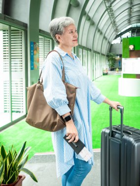 Yaşlı, yaşlı Asyalı bir kadının portresi. 60 'lı yaşlarda bavul çantasıyla gülümsüyor ve seyahat konsepti için pasaport tutuyor..
