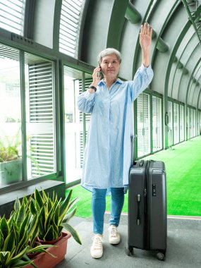 Yaşlı, yaşlı Asyalı bir kadının portresi. 60 'lı yaşlarda bavul çantasıyla gülümsüyor ve seyahat konsepti olarak akıllı telefon kullanıyor..