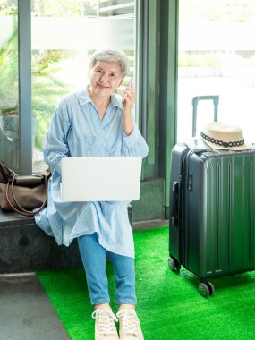 Rahatla, tatil, seyahat, yaşlı Asyalı bir kadının portresi 60 'lı yaşlarda bilgisayarlı dizüstü bilgisayar ve akıllı telefon kullanarak seyahat zamanlarını beklerken gülümsüyor..