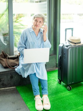 Rahatla, tatil, seyahat, yaşlı Asyalı bir kadının portresi 60 'lı yaşlarda bilgisayarlı dizüstü bilgisayar ve akıllı telefon kullanarak seyahat zamanlarını beklerken gülümsüyor..