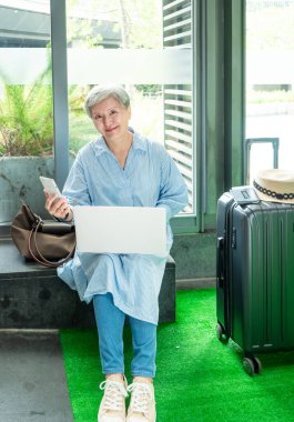 Rahatla, tatil, seyahat, yaşlı Asyalı bir kadının portresi 60 'lı yaşlarda bilgisayarlı dizüstü bilgisayar ve akıllı telefon kullanarak seyahat zamanlarını beklerken gülümsüyor..