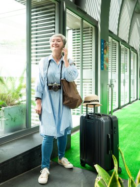 Yaşlı, yaşlı Asyalı bir kadının portresi. 60 'lı yaşlarda bavul çantasıyla gülümsüyor ve seyahat konsepti olarak akıllı telefon kullanıyor..