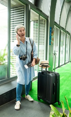 Yaşlı, yaşlı Asyalı bir kadının portresi. 60 'lı yaşlarda bavul çantasıyla gülümsüyor ve seyahat konsepti olarak akıllı telefon kullanıyor..