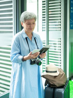 Yaşlı, yaşlı Asyalı bir kadının portresi. 60 'lı yaşlarda bavul çantasıyla gülümsüyor ve seyahat konsepti olarak akıllı telefon kullanıyor..