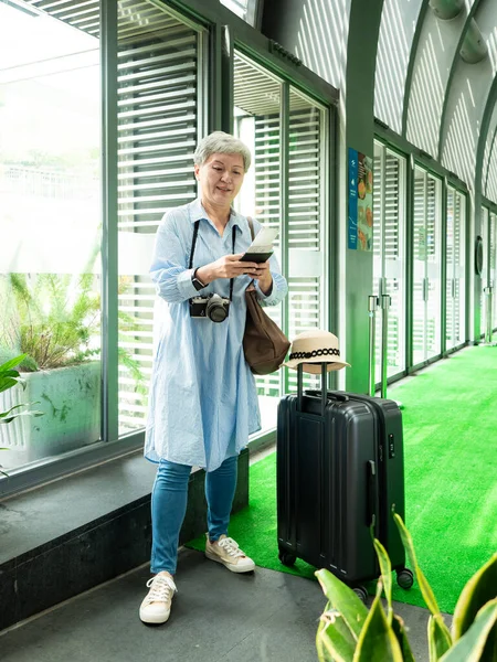 Yaşlı, yaşlı Asyalı bir kadının portresi. 60 'lı yaşlarda bavul çantasıyla gülümsüyor ve seyahat konsepti olarak akıllı telefon kullanıyor..