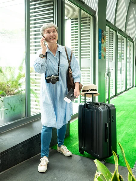 Yaşlı, yaşlı Asyalı bir kadının portresi. 60 'lı yaşlarda bavul çantasıyla gülümsüyor ve seyahat konsepti olarak akıllı telefon kullanıyor..