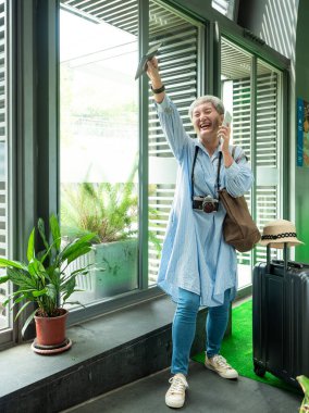 Yaşlı, yaşlı Asyalı bir kadının portresi. 60 'lı yaşlarda bavul çantasıyla gülümsüyor ve seyahat konsepti olarak akıllı telefon kullanıyor..