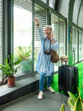 Yaşlı, yaşlı Asyalı bir kadının portresi. 60 'lı yaşlarda bavul çantasıyla gülümsüyor ve seyahat konsepti olarak akıllı telefon kullanıyor..