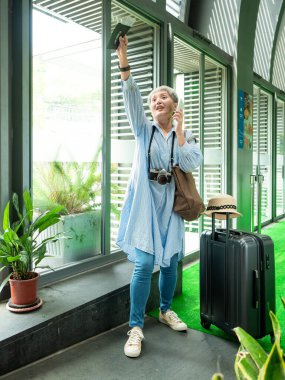 Yaşlı, yaşlı Asyalı bir kadının portresi. 60 'lı yaşlarda bavul çantasıyla gülümsüyor ve seyahat konsepti olarak akıllı telefon kullanıyor..