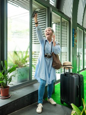 Yaşlı, yaşlı Asyalı bir kadının portresi. 60 'lı yaşlarda bavul çantasıyla gülümsüyor ve seyahat konsepti olarak akıllı telefon kullanıyor..