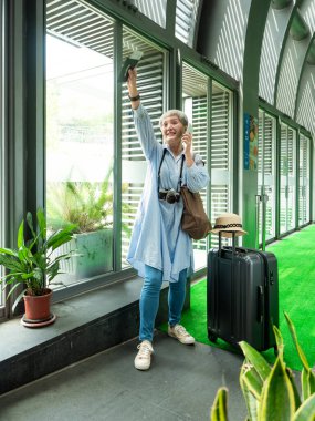 Yaşlı, yaşlı Asyalı bir kadının portresi. 60 'lı yaşlarda bavul çantasıyla gülümsüyor ve seyahat konsepti olarak akıllı telefon kullanıyor..