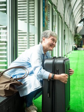 Yaşlı, yaşlı Asyalı bir kadının portresi. 60 'larında gülümseyen, bavul çantasıyla oturan ve seyahat konsepti için pasaport taşıyan..