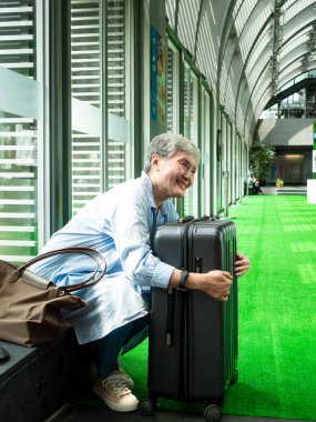Yaşlı, yaşlı Asyalı bir kadının portresi. 60 'larında gülümseyen, bavul çantasıyla oturan ve seyahat konsepti için pasaport taşıyan..