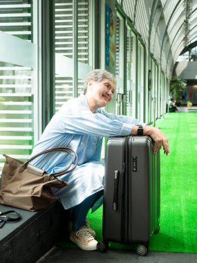 Yaşlı, yaşlı Asyalı bir kadının portresi. 60 'larında gülümseyen, bavul çantasıyla oturan ve seyahat konsepti için pasaport taşıyan..
