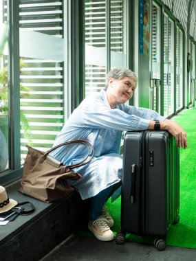 Yaşlı, yaşlı Asyalı bir kadının portresi. 60 'larında gülümseyen, bavul çantasıyla oturan ve seyahat konsepti için pasaport taşıyan..