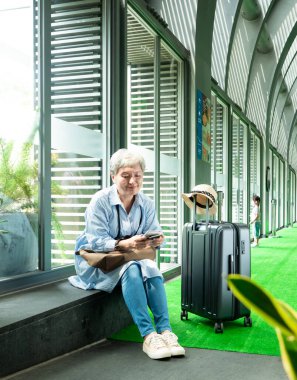 Yaşlı, yaşlı Asyalı bir kadının portresi. 60 'larında gülümseyen, bavul çantasıyla oturan ve seyahat konsepti için pasaport taşıyan..