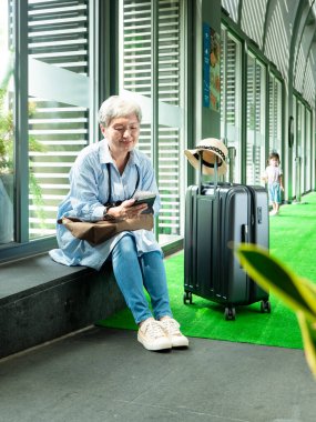 Yaşlı, yaşlı Asyalı bir kadının portresi. 60 'larında gülümseyen, bavul çantasıyla oturan ve seyahat konsepti için pasaport taşıyan..