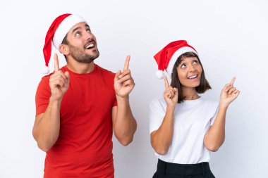 Beyaz arka planda izole edilmiş Noel şapkalı genç çift işaret parmağıyla işaret etmek harika bir fikir.