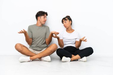 Beyaz arka planda izole bir şekilde yerde oturan genç melez çift omuzlarını kaldırırken önemsiz bir jest yapıyor.