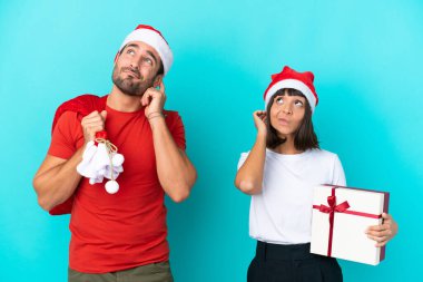 Noel şapkalı genç bir çift mavi arka planda izole edilmiş hediyeler dağıtıyor.