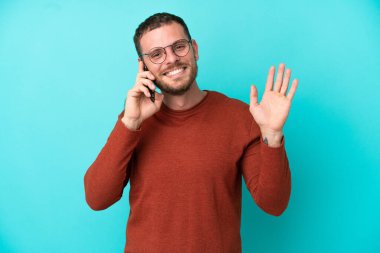 Mavi arka planda izole edilmiş cep telefonu kullanan Brezilyalı genç adam mutlu bir ifadeyle selamlıyor.