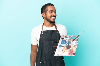 Mavi arka planda izole edilmiş bir palet tutan genç sanatçı yana bakıyor ve gülümsüyor.