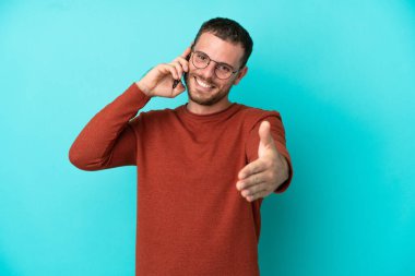 Mavi arka planda izole edilmiş cep telefonu kullanan genç Brezilyalı adam iyi bir anlaşma için el sıkışıyor.