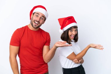 Beyaz arka planda izole edilmiş Noel şapkalı genç çift, davet edilmeleri için ellerini yana uzatıyor.