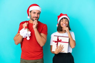 Noel şapkalı genç bir çift mavi arka planda izole edilmiş hediyeler dağıtıyor tatlı bir ifadeyle gülümsüyor.