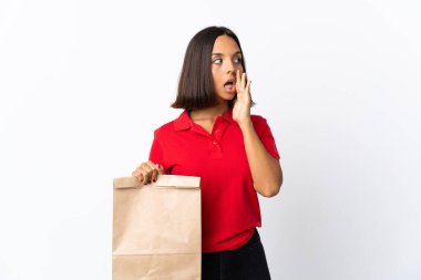 Beyaz arka planda izole edilmiş alışveriş torbası tutan genç Latin kadın yan tarafa bakarken sürpriz bir jest yaparak bir şeyler fısıldıyor.