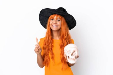 Cadı kılığında genç bir kadın beyaz arka planda izole edilmiş bir kafatası tutuyor ve parmağını kaldırıyor.