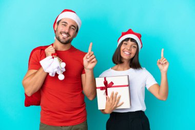 Noel şapkalı genç çift mavi arka planda izole edilmiş hediyeler dağıtıyor ve en iyinin işareti olarak parmağını kaldırıyor.