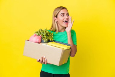 Genç Uruguaylı kız sarı arka planda izole edilmiş şeylerle dolu bir kutuyu kaldırırken hareket ediyor ve ağzı sonuna kadar açık bağırıyor.