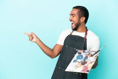 Mavi arka planda izole edilmiş bir palet tutan genç sanatçı parmağını yana uzattı ve bir ürün sundu.
