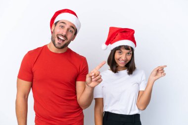 Beyaz arka planda izole edilmiş Noel şapkalı genç bir çift parmak göstererek bir ürün sunuyor.