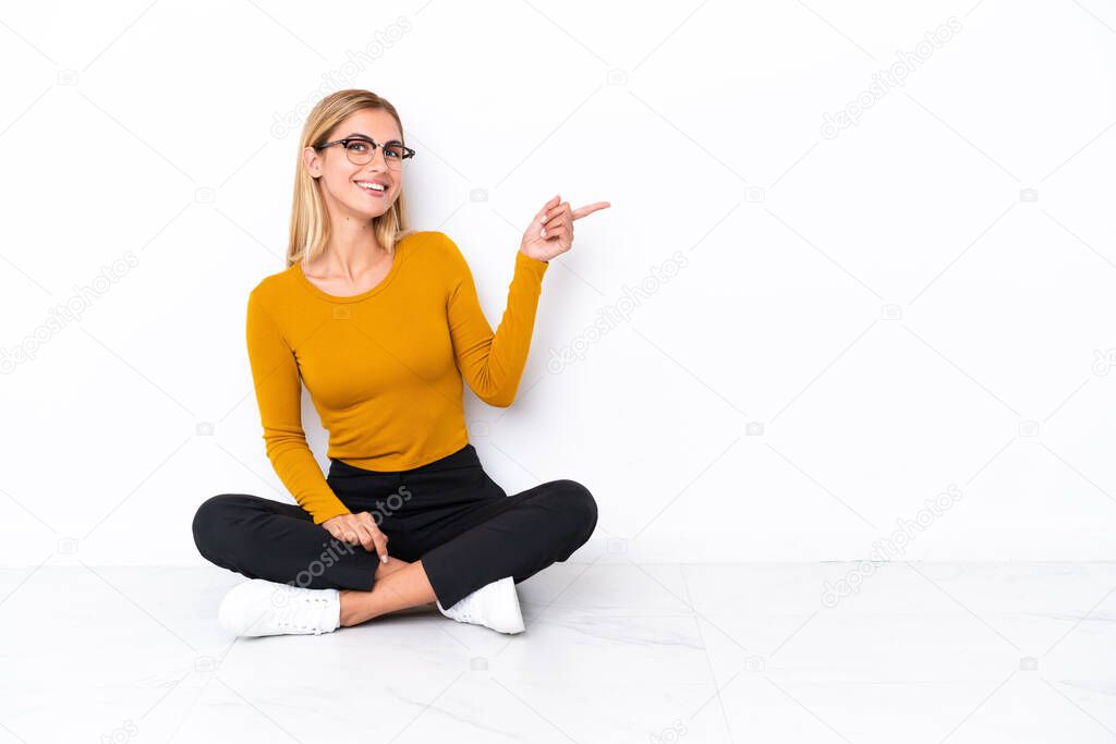 Blonde Uruguayan girl sitting on the floor pointing finger to the side