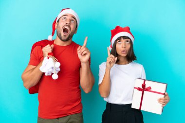 Noel şapkalı genç bir çift mavi arka planda izole edilmiş hediyeler dağıtıyor.