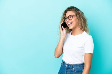Kıvırcık saçlı kız mavi arka planda izole edilmiş cep telefonuyla sohbet ediyor.
